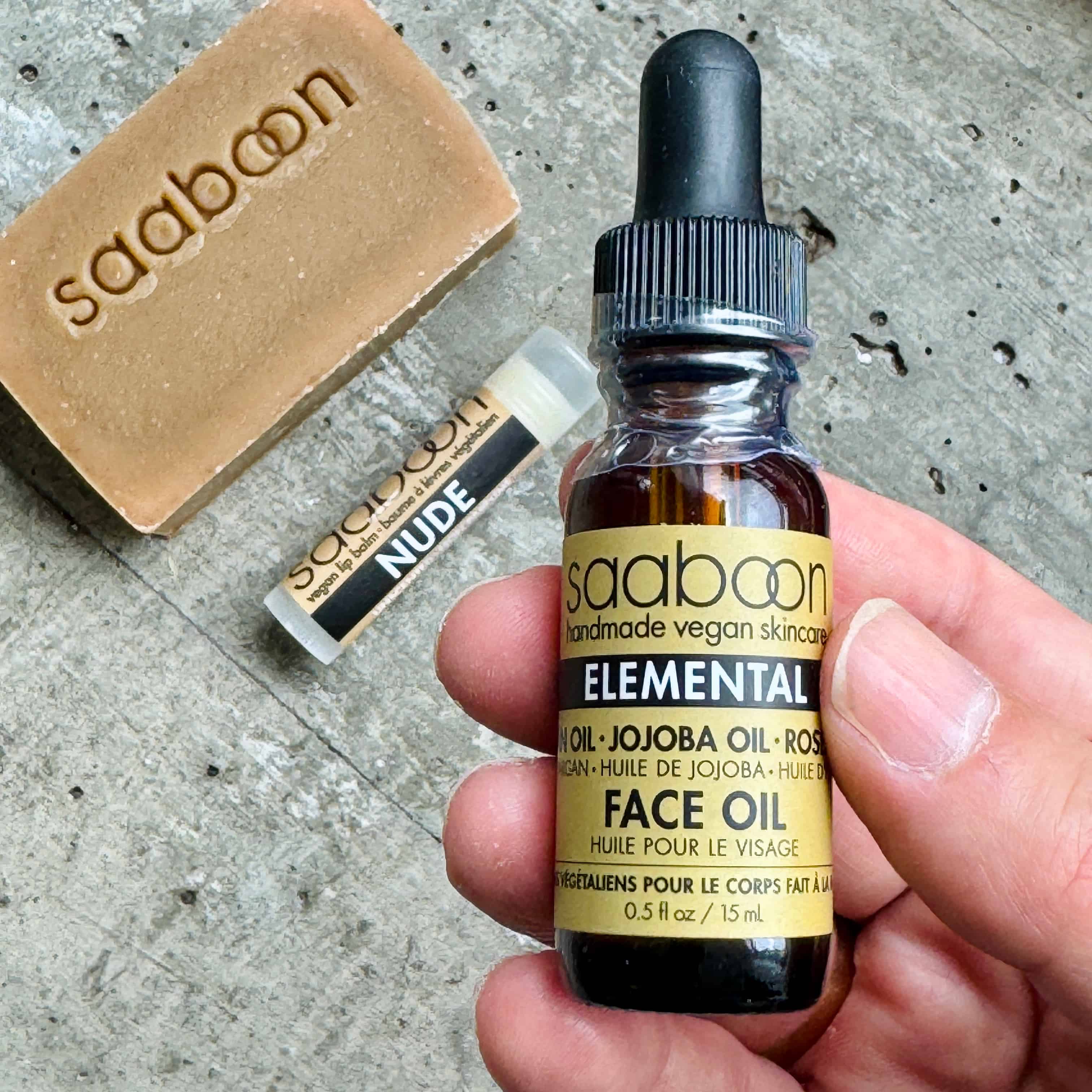 A hand holds a small glass dropper bottle of saaboon Elemental Face Oil next to a bar of saaboon soap and a lip balm tube on a textured gray surface.