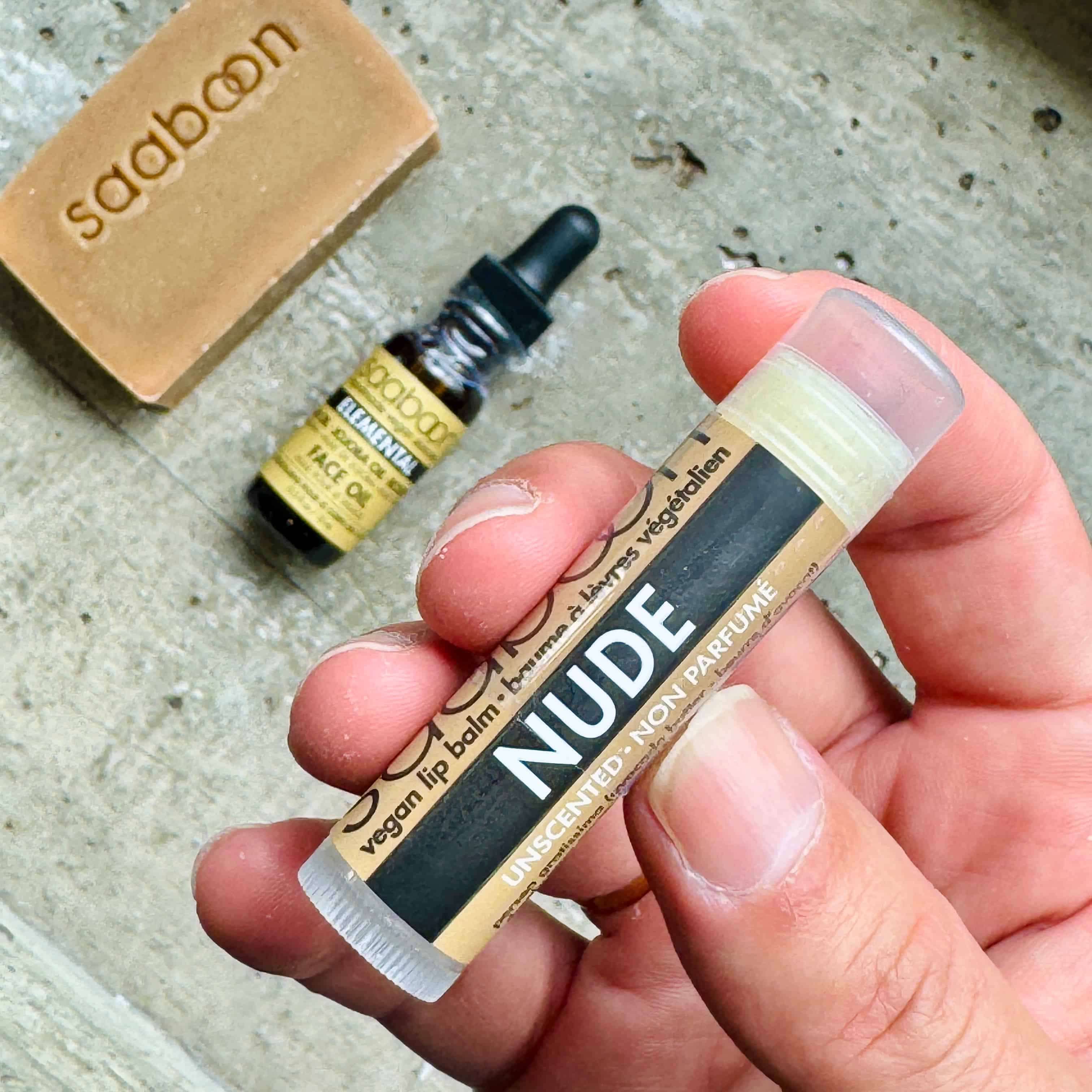 A hand holds a tube of nude-colored vegan lip balm with a black and beige label that reads 'NUDE'. In the background, a small bottle of soap and a bar of soap engraved with 'saaboon' are visible on a textured gray surface.