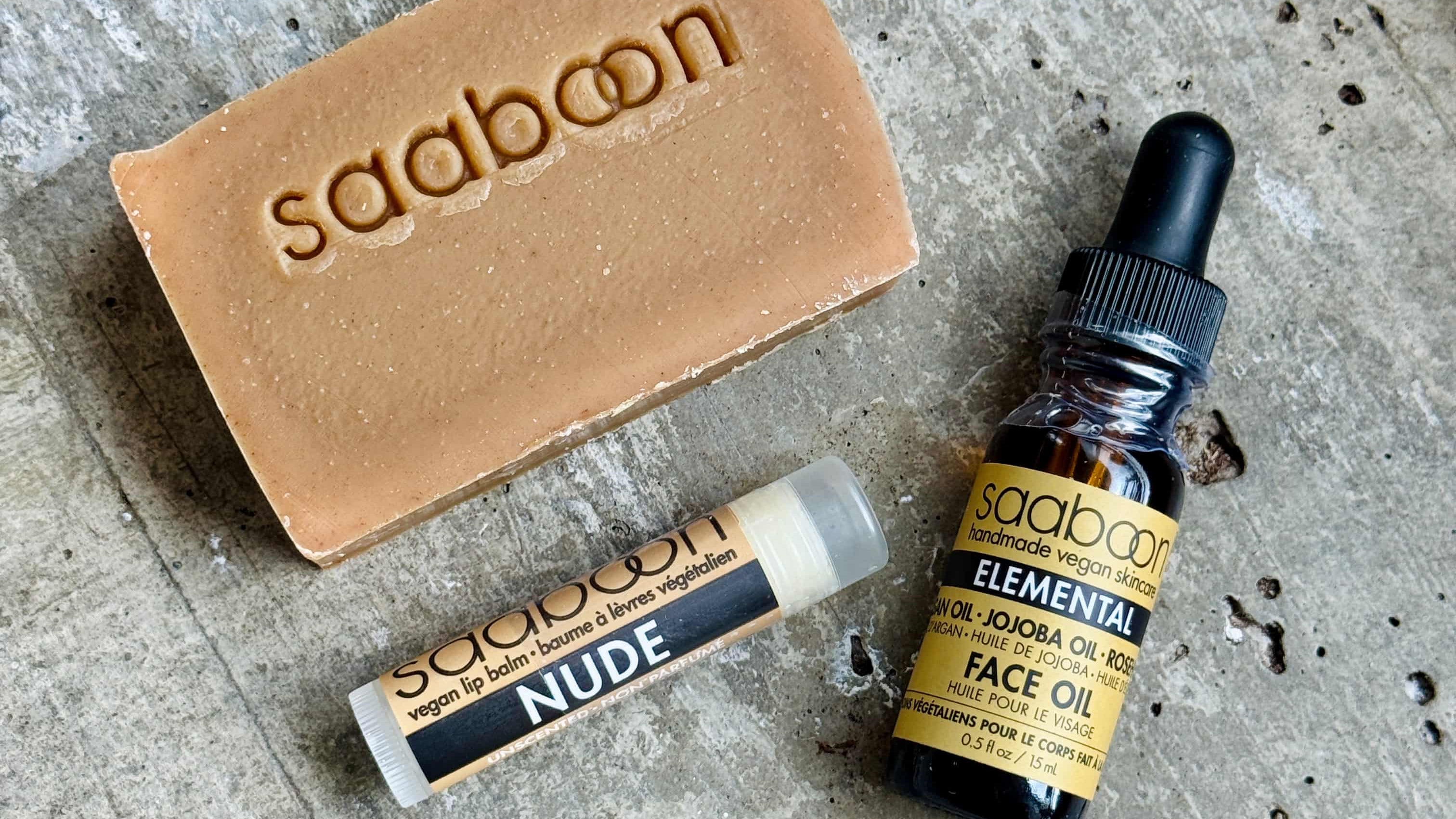 Three Saaboon skincare products arranged on a textured gray stone surface: a tan soap bar with the brand name embossed, a small lip balm tube labeled NUDE, and a dark amber face oil bottle with a black dropper cap and yellow label.