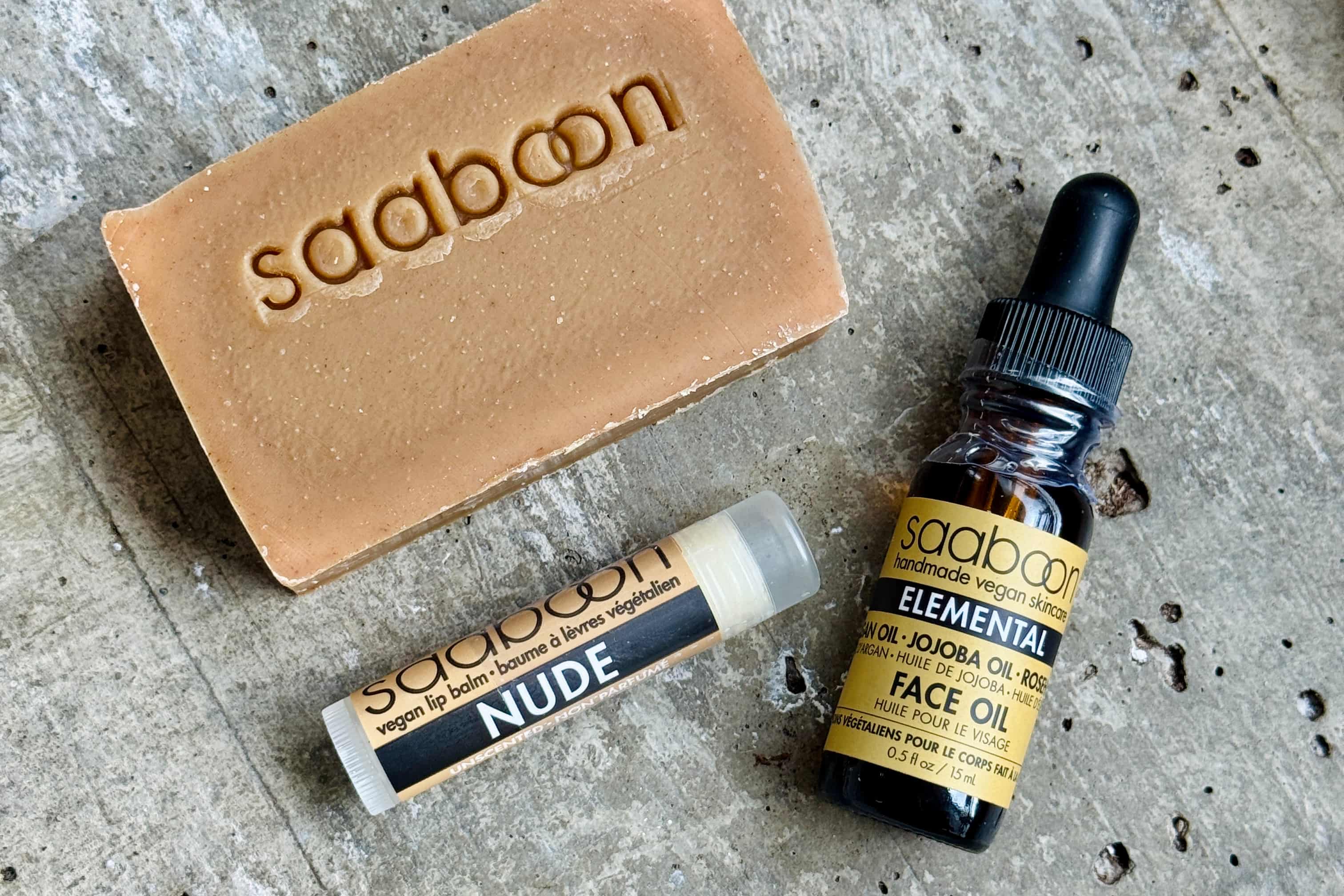 Three Saaboon skincare products arranged on a textured gray stone surface: a tan soap bar with the brand name embossed, a small lip balm tube labeled NUDE, and a dark amber face oil bottle with a black dropper cap and yellow label.
