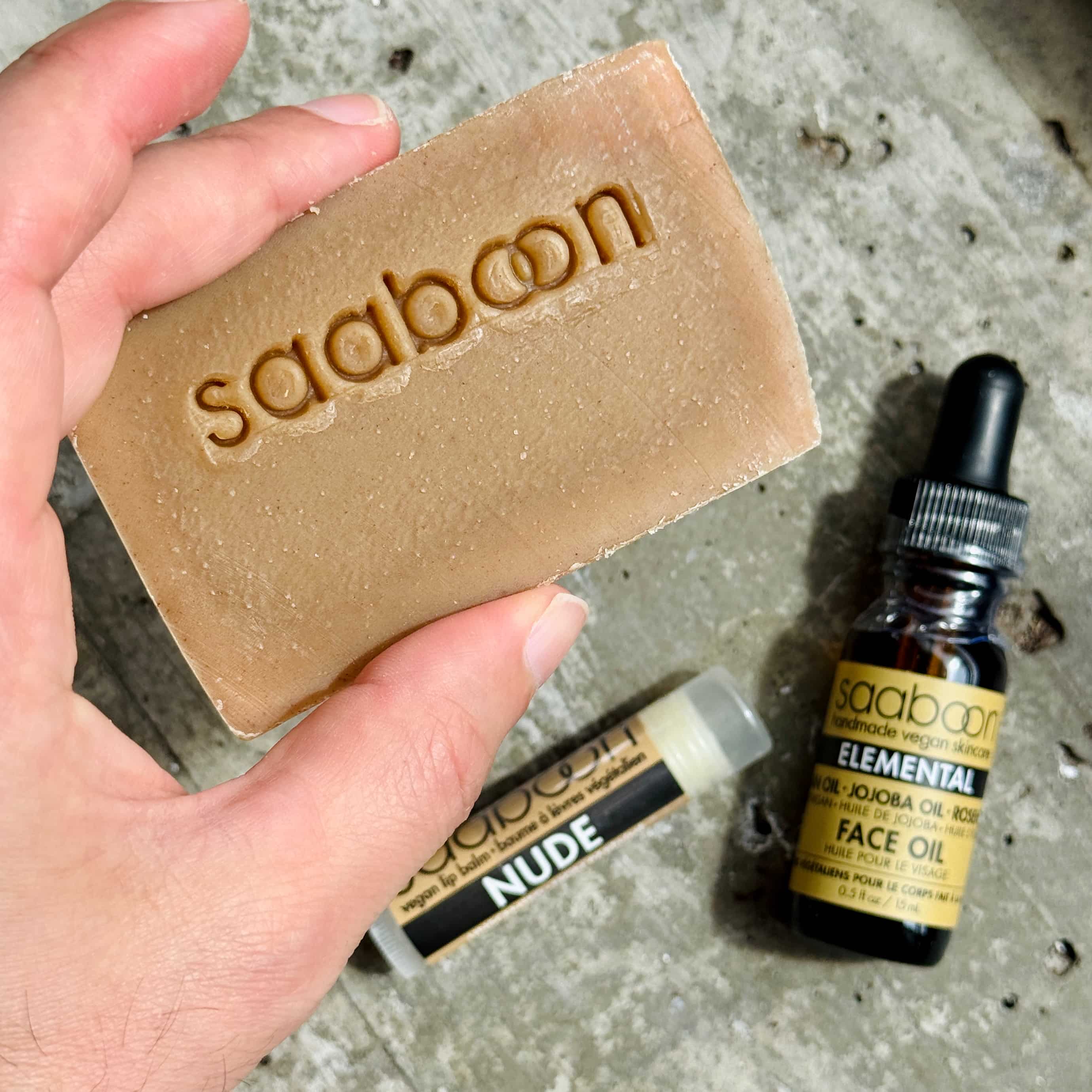 A hand holds a rectangular bar of soap embossed with the brand name 'saaboon' in a natural tan shade. In the background on a textured gray surface, a small amber bottle of saaboon Elemental Face Oil and a lip balm tube labeled NUDE are visible.