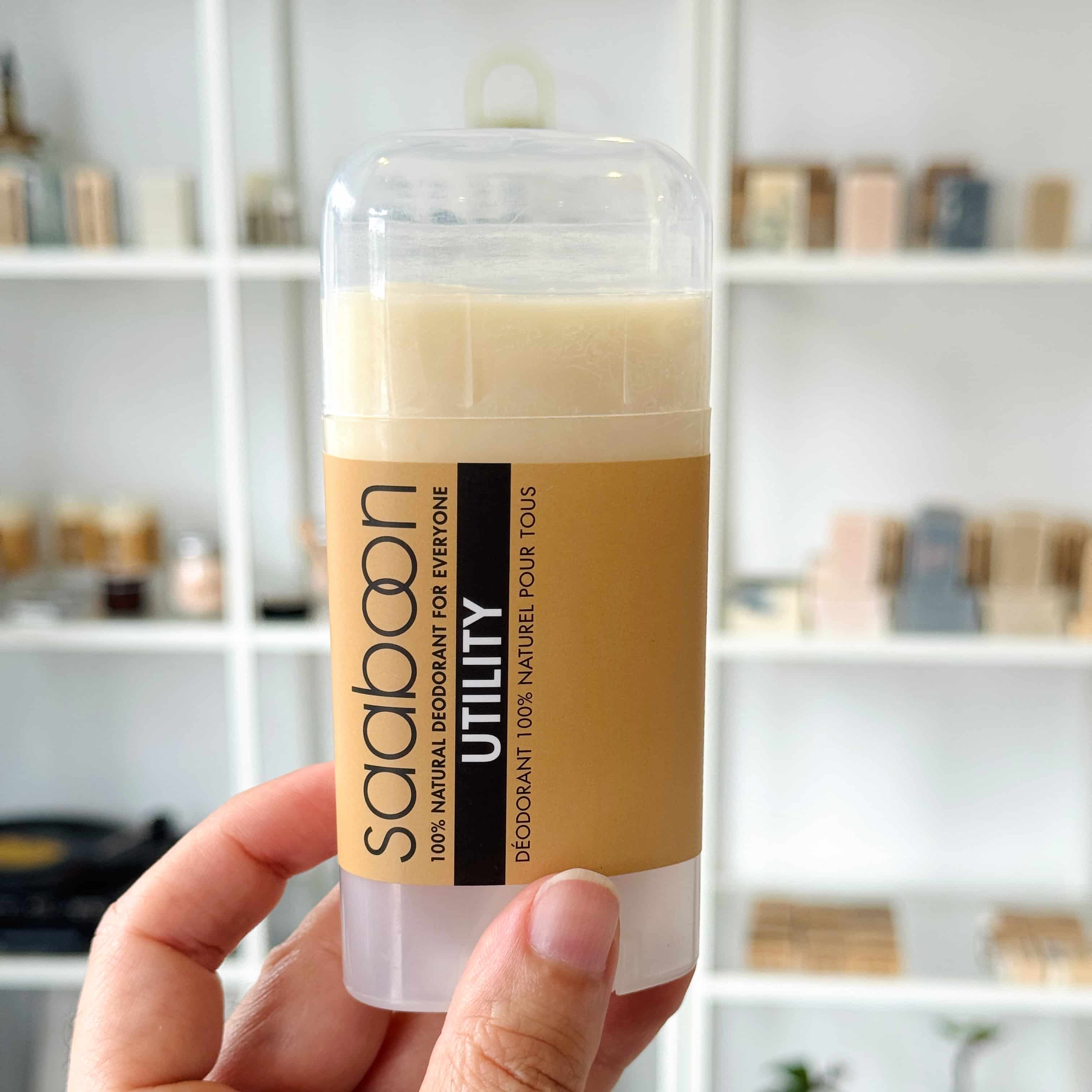 A hand holding a stick deodorant labeled saaboon UTILITY against a blurred saaboon shop background of white shelves with decorative items.