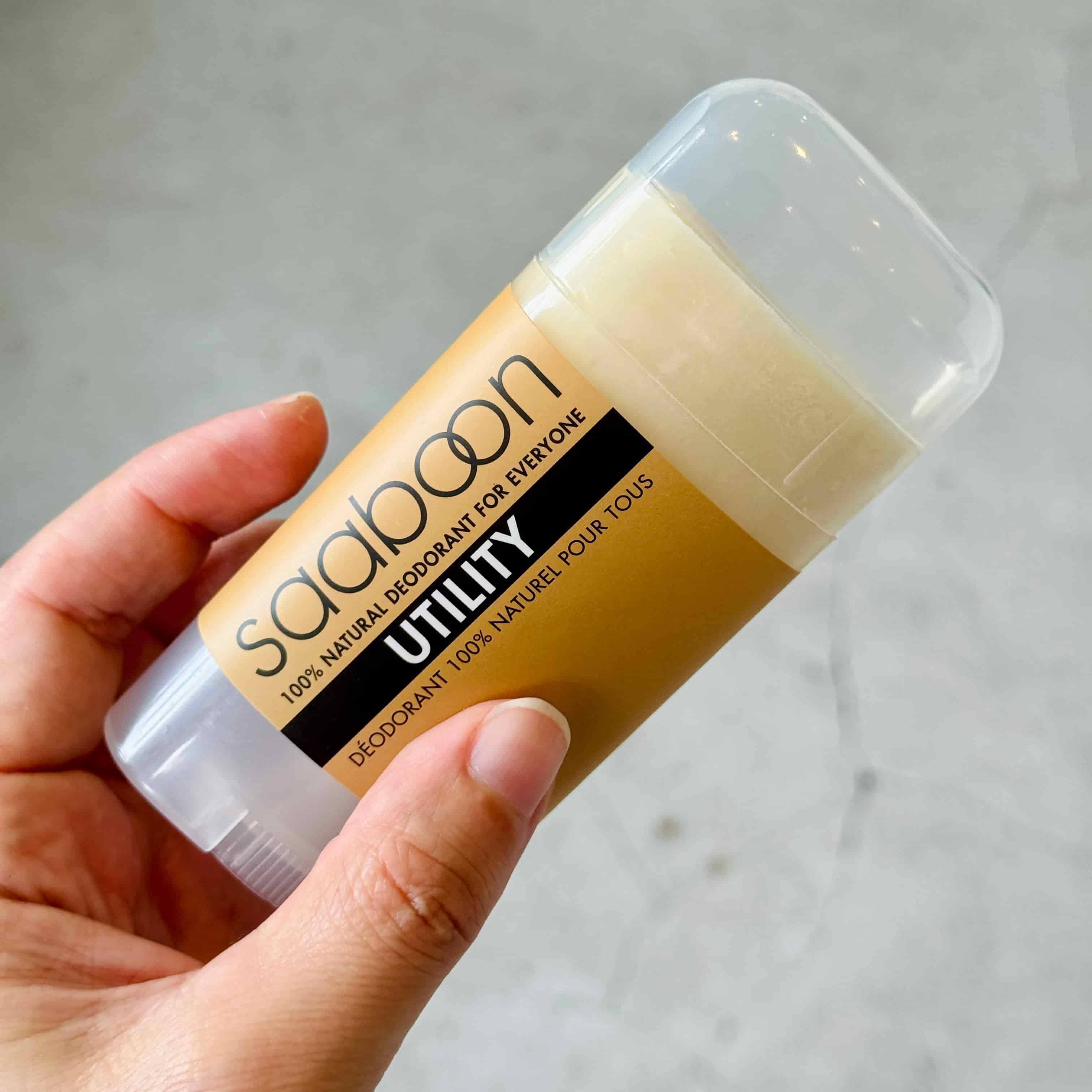 A hand holding a Saaboon Utility deodorant stick against a light gray stone background.
