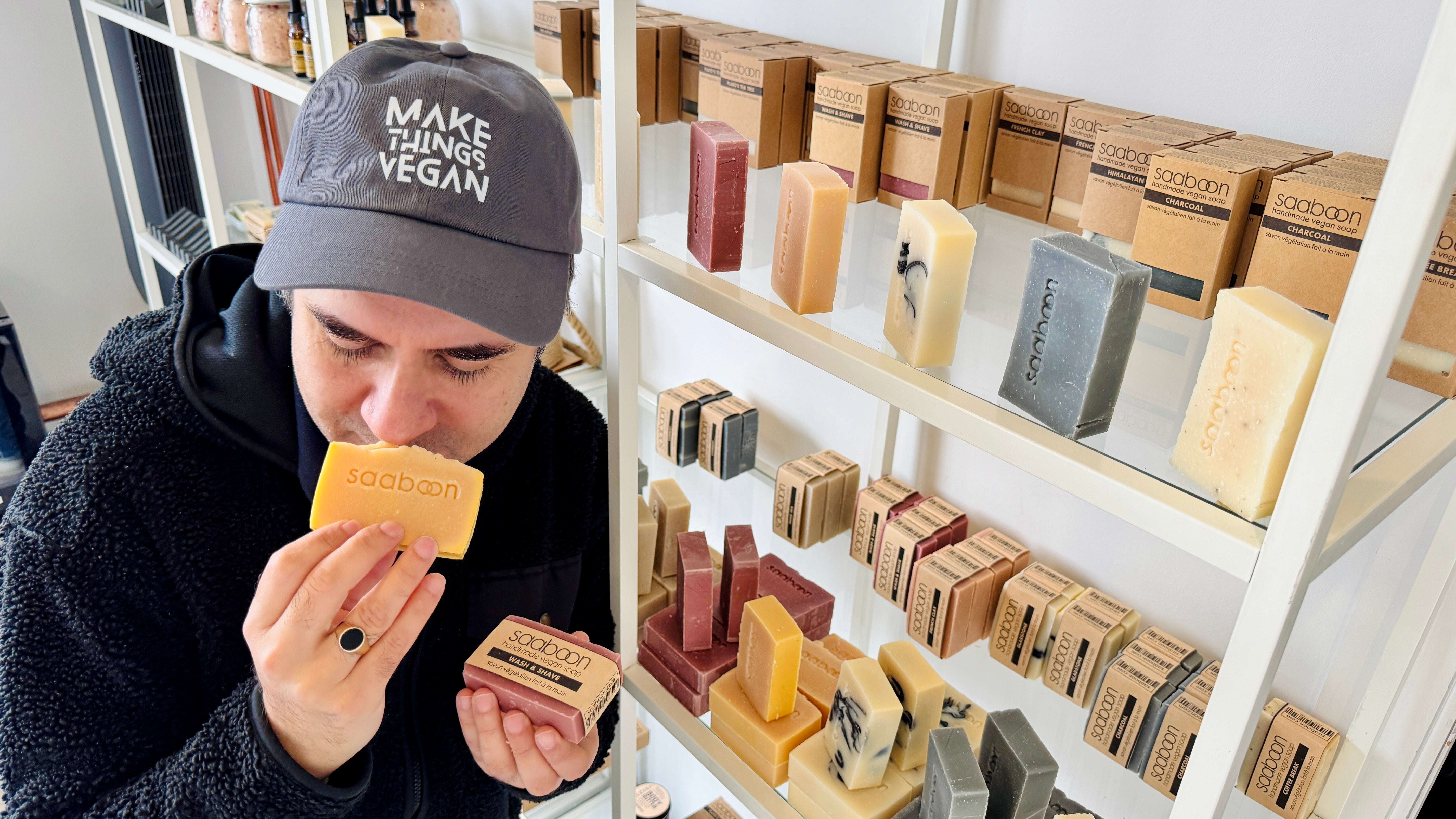 Person smelling a bar of soap in front of a display of various soaps, wearing a hat reading Make Things Vegan.