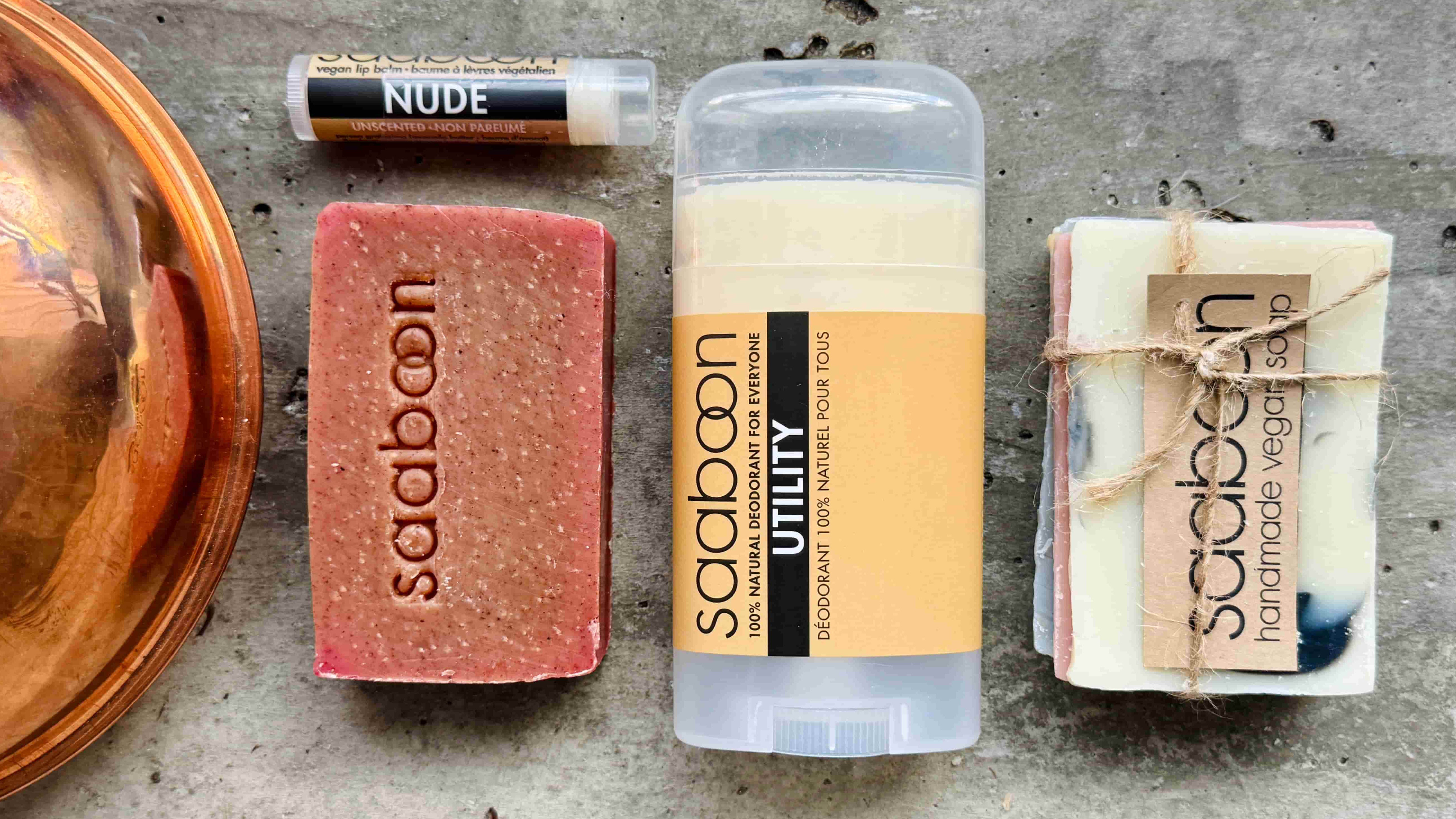 A flat-lay of three Saaboon natural soap products on a gray concrete surface: a pinkish-red soap bar, a clear stick container with a yellow label, and a wrapped white marbled soap bar tied with twine, alongside a copper bowl edge and a lip balm tube labeled NUDE.