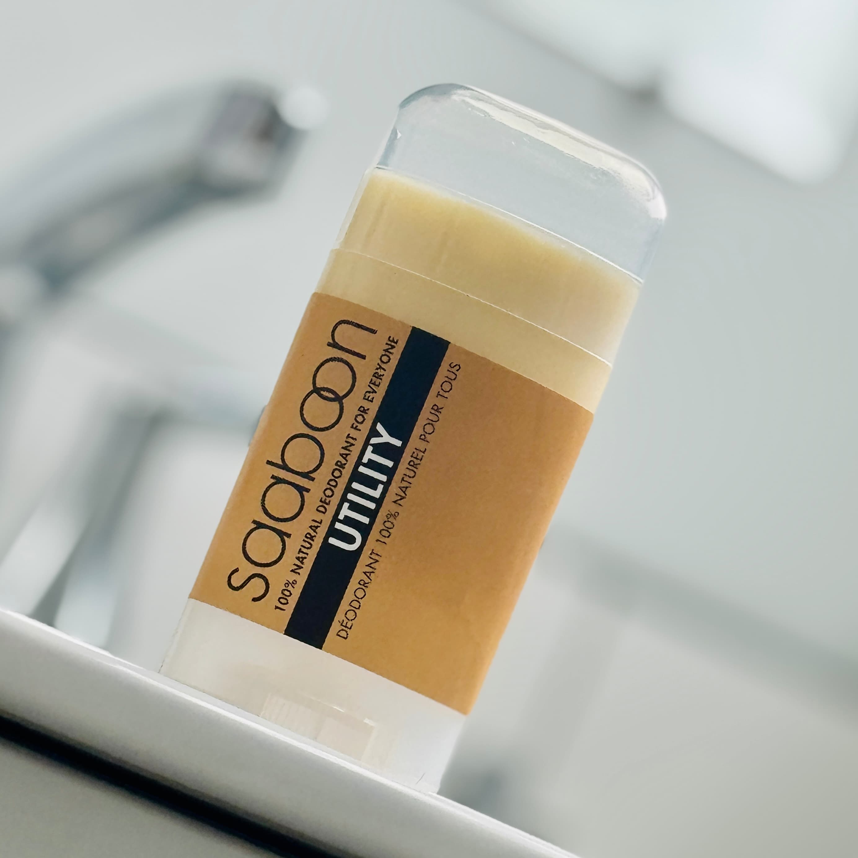 A close-up of a Saaboon Utility deodorant stick in a clear container with a Kraft and black label, resting on a white surface with a blurred bathroom sink and faucet in the background.